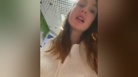 Media: A blurry video of a young Caucasian woman with fair skin and long, auburn hair, wearing a white, ribbed sweater, captured from a low angle, indoors, with a white grid-patterned wall and green foliage in the background.