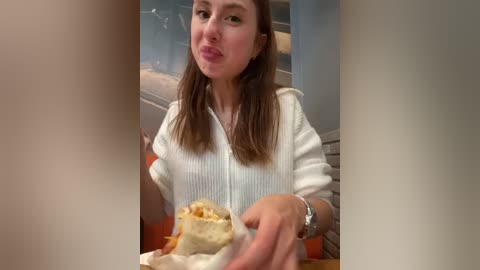 Media: Video of a young woman with light skin and long brown hair, wearing a white knit cardigan, blowing a kiss while holding a wrapped burrito in a casual indoor setting.