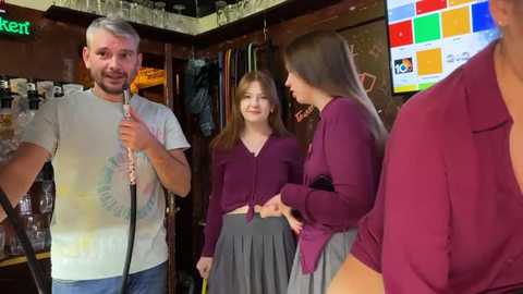 Media: Video of a casual, indoor setting featuring three young women in maroon tops, one holding a feather duster, another wearing a gray skirt. Background includes a bar with shelves and a TV screen.