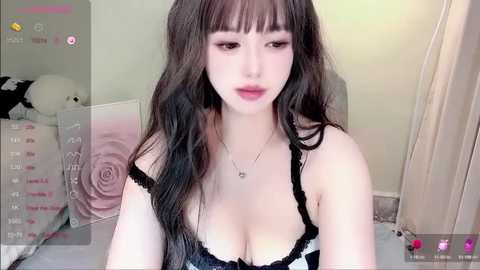 Video of an East Asian woman with long, wavy black hair, fair skin, wearing a black lace bralette, in a softly lit bedroom.