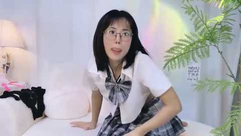 Media: A video of an Asian woman with straight black hair, glasses, and a white plaid schoolgirl outfit, posing seductively on a white couch in a modern, brightly lit room with greenery.