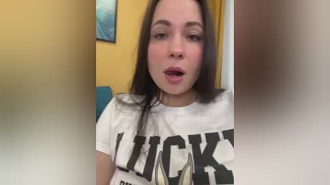 Media: Video of a fair-skinned, brunette woman with straight hair, wearing a white T-shirt with \"LUCK\" in large black letters. She has a neutral expression and is indoors with yellow walls and a blue piece of furniture in the background.