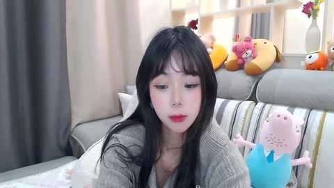 Media: Video of an Asian woman with long black hair and red lipstick, sitting on a striped sofa, surrounded by plush toys in a cozy, sunlit room.