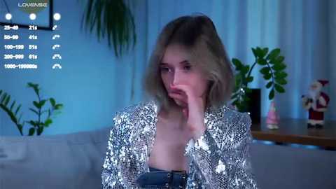 Media: Video of a young woman with shoulder-length blonde hair, wearing a silver sequined jacket and blue top, looking contemplative in a modern living room with plants, a Christmas tree, and a TV screen displaying live camera feed.