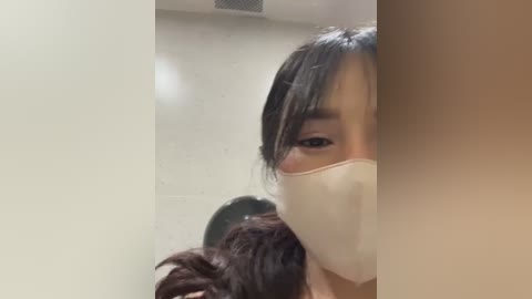 Media: Video of an East Asian woman with medium skin tone, long black hair, and a beige face mask, partially obscured by a black-haired woman with a dark complexion in the background.