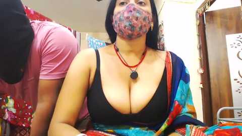 Media: Video of a dark-skinned woman with large breasts wearing a black bra, colorful saree, and a face mask, standing in a crowded, brightly lit room with wooden furniture and people in the background.