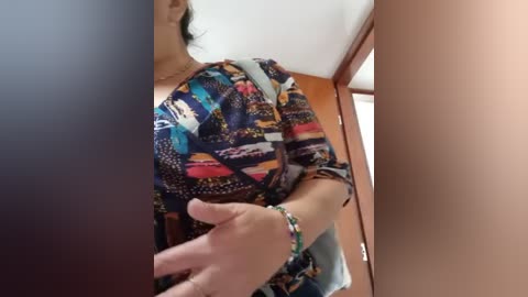 Media: Video of a woman with medium skin tone, wearing a colorful, patterned blouse, and a green bracelet. She stands in a room with wooden furniture and a mirror.