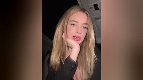 Media: Video of a young blonde woman with fair skin, wearing a black top, resting her chin on her hand, with a dimly lit background showing part of a car interior.