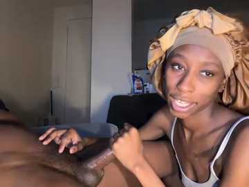 Media: Video of a young Black woman with a light brown headscarf, smiling, holding an erect, circumcised penis, in a dimly lit bedroom.