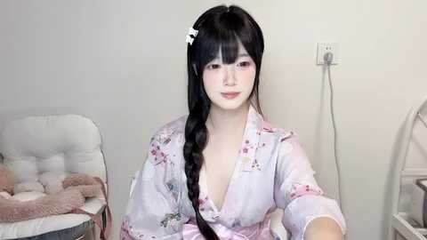 Media: Video of an East Asian woman with long black hair in a braid, wearing a light pink floral blouse, in a simple room with a white chair and a beige blanket.