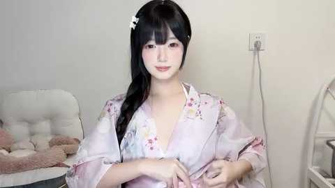 Media: Video of a young Asian woman with long black hair, wearing a light pink kimono with floral patterns, sitting in a cozy, softly lit room with a white chair and a visible electrical outlet.