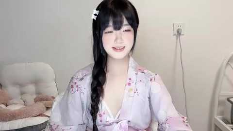 Media: Video of a young Asian woman with long black hair, wearing a light pink floral kimono, sitting in a cozy, dimly lit room with a plush white chair and a white power outlet in the background.