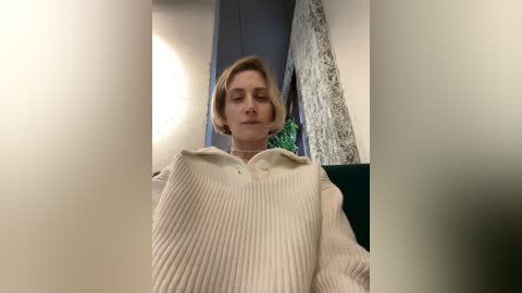 Video of a blonde woman with short hair, wearing a beige ribbed sweater, seated indoors with a white wall and green leafy plant in the background.