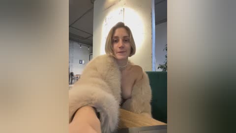 Media: Video of a blonde woman with a small breast size, wearing a fur coat and sitting in a modern, dimly lit cafe.