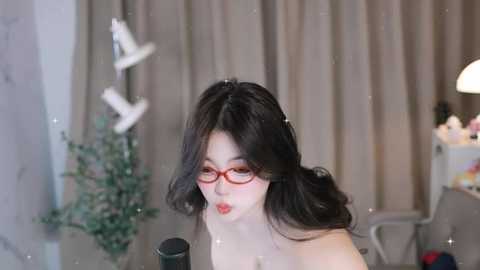 Media: Video of an Asian woman with long black hair, red glasses, and fair skin, puckering her lips in a suggestive manner. Background features a green Christmas tree, beige curtains, and a modern desk with a lamp.