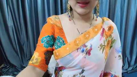 Media: Video of a light-skinned woman wearing an orange saree with blue floral embroidery and a white floral blouse. She has dangling earrings and a necklace, with a gray, draped background.