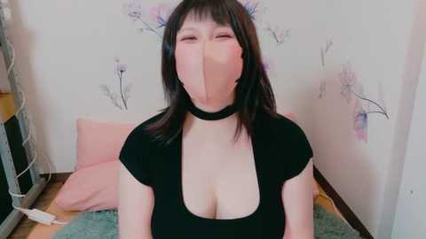 Media: Video of an East Asian woman with black hair and bangs, wearing a black choker and crop top, with a pink mask covering her nose and mouth. She sits on a pink cushion in a room with floral wallpaper and a wooden floor.