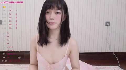 Media: Video of a young Asian woman with straight black hair and light skin, wearing a pink lace camisole, sitting on a wooden floor, calendar visible in the background.