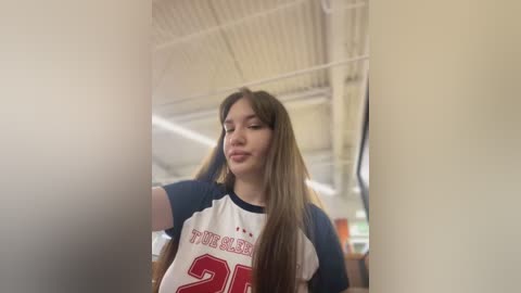 Media: Video of a young woman with long brown hair, wearing a white and blue \"Purdue\" jersey, standing indoors with a gym's high ceiling and bright lights in the background.