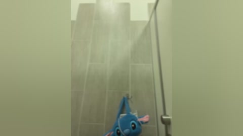 Media: Video of a blue, cartoonish character with large eyes and ears, standing under a shower with water cascading down, in a dimly lit, minimalist bathroom.