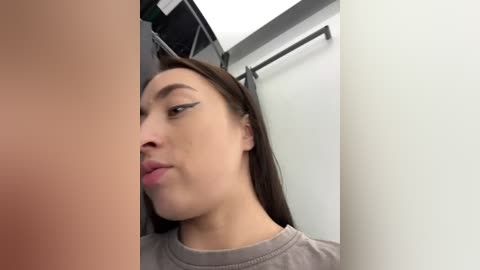 Media: Video of a young Asian woman with light skin, brown hair, and black eyeliner, puckering her lips in a bathroom stall with a beige curtain.