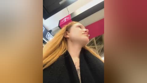 Media: Video of a young Caucasian woman with long, straight, light brown hair, wearing a black coat and a pink hat. Background shows a modern, indoor setting with striped walls and an abstract light fixture.