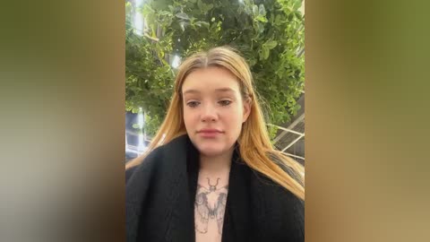 Media: Video of a young woman with long blonde hair, wearing a black coat, standing in front of a lush green tree, showcasing a detailed deer tattoo on her chest.