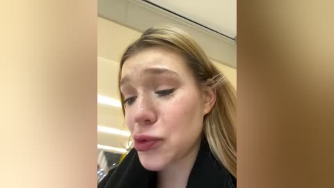 Media: Video of a young woman with fair skin, long blonde hair, and a black top, making a silly face in a bathroom with beige walls and fluorescent lights.