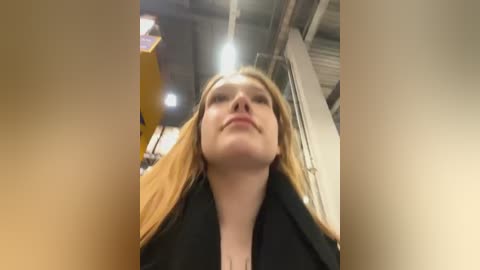 Media: Video of a young woman with fair skin and long blonde hair, wearing a black top, standing in a brightly lit indoor space with visible industrial ceiling beams and yellow and white structures in the background.