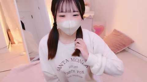 Media: A video of a young East Asian woman with straight black hair in pigtails, wearing a white sweatshirt with text, a white mask, and a pink heart pillow. She stands indoors in a softly lit room with beige carpeting and a mirror.