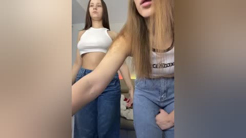 Media: Video of two young women in casual attire, one in a white crop top and high-waisted jeans, the other in a cropped white top and denim shorts, standing in a modern living room.