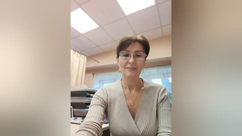 Media: Video of a middle-aged woman with short brown hair, wearing glasses and a beige V-neck sweater, standing in a modern medical office with fluorescent lighting, white walls, and medical equipment in the background.