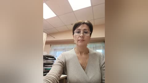 Media: A video of a middle-aged woman with short brown hair, wearing glasses and a beige ribbed V-neck sweater, standing in a beige office with fluorescent lights, stacked papers, and a window.