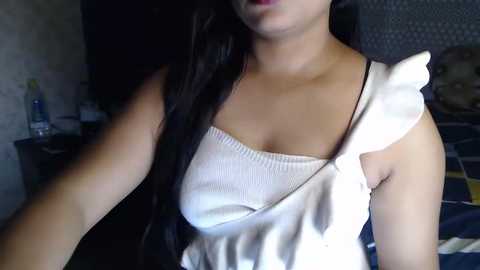 Media: A video of a young woman with long black hair, wearing a white, sleeveless, ribbed top, sitting in a dimly lit room with a dark gray headrest and a partially visible bed with dark bedding.