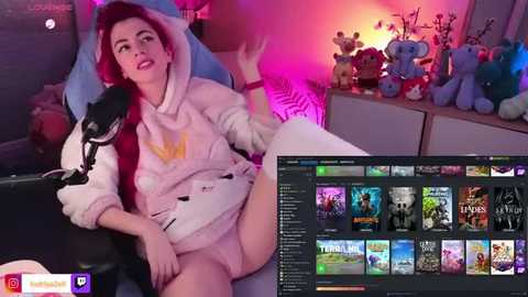 Media: A video of a pale-skinned woman with red hair wearing a white robe, sitting on a gaming chair with a headset, surrounded by plush toys and a TV screen displaying a gaming interface.