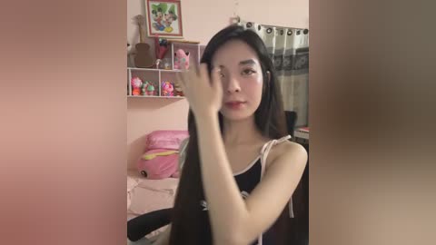 Media: A video of an East Asian woman with long black hair, wearing a black sleeveless top, adjusting her hair in a pink room with shelves and toys.