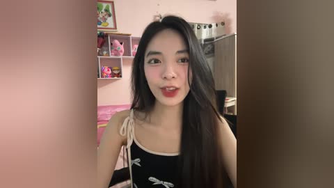 Media: Video of an East Asian woman with long black hair, wearing a black tank top with white bow accents. Background includes pink walls, stuffed animals, and shelves.