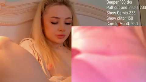 Media: A close-up video of a young, fair-skinned woman with blonde hair, wearing a white shirt, lying on a bed, with a pink, soft-focus background.