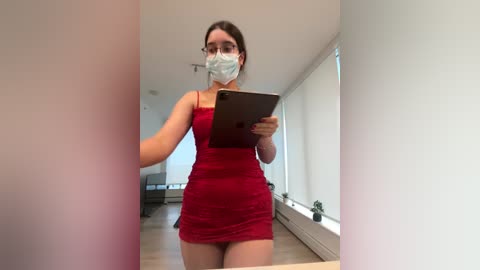 A video of a woman with light skin, dark hair, and a slim build wearing a tight red dress, a face mask, and glasses, holding a clipboard, standing in a minimalist, brightly lit room with white walls and large windows.