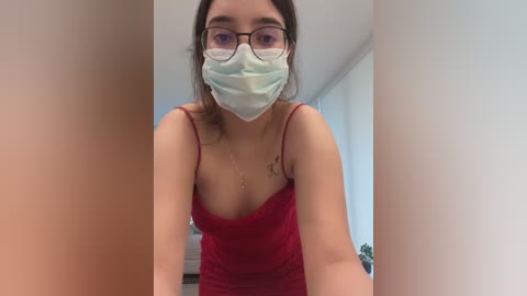 Media: A video of a woman with fair skin, dark hair, and glasses, wearing a red tank top, mask, and necklace. The image is blurry, focusing on her breasts.