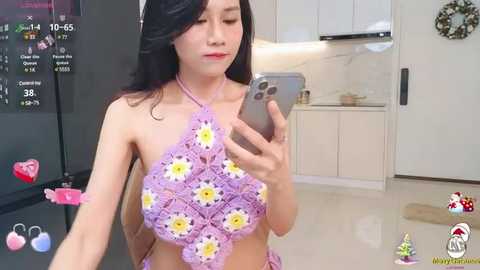 Media: A young Asian woman with long black hair and fair skin takes a selfie in a modern kitchen, wearing a pink crochet bikini with flower embellishments.