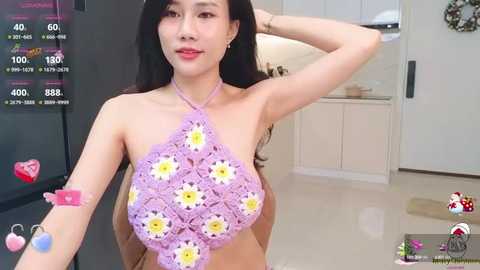 Media: Video of an Asian woman with long black hair, fair skin, and a slender build, wearing a pastel purple crochet halter top adorned with large yellow daisies. She stands in a modern, brightly lit kitchen with white cabinets and a stainless steel fridge.