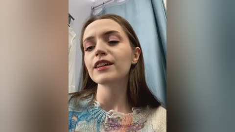 Media: Video of a young woman with fair skin, brown hair, and pink makeup, wearing a colorful, textured sweater, standing in a bathroom with light blue shower curtain.