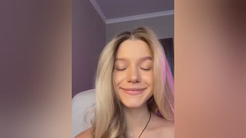 Media: Video of a smiling, fair-skinned blonde woman with closed eyes, wearing a necklace, in a room with white walls and a pink curtain.