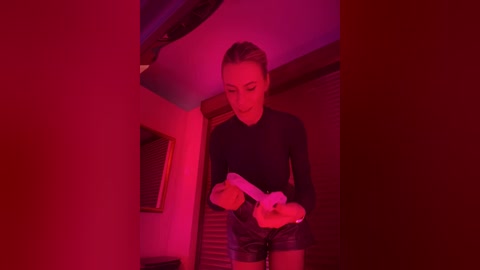 Video of a slender woman in a black top, holding a white remote, standing in a dimly lit room with red lighting.