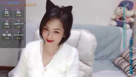 Media: A video of a smiling East Asian woman with black cat ears, red lipstick, and a white robe, sitting on a light blue couch, with a stuffed animal and a digital display in the background.