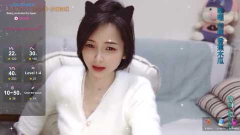 Media: Video of an East Asian woman with short black hair, wearing a white cardigan, seated on a couch. Background shows a gaming interface with virtual cat ears.