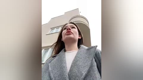 Media: Video of a young woman with fair skin and straight brown hair, wearing a gray coat over a white shirt, standing in front of a beige building. The image is shot from a low angle, emphasizing her height.