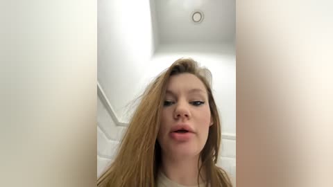 Media: Video of a young Caucasian woman with long, straight, light brown hair, light skin, and a neutral expression. She's standing in a narrow, bright white hallway with a ceiling light visible.