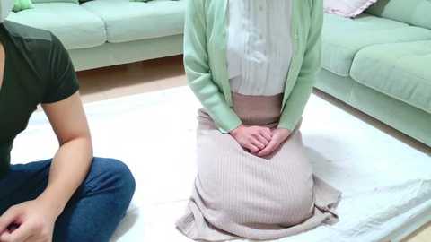 Media: Video of a person kneeling on a light-colored rug in a living room, wearing a white blouse, pastel green cardigan, and beige skirt.